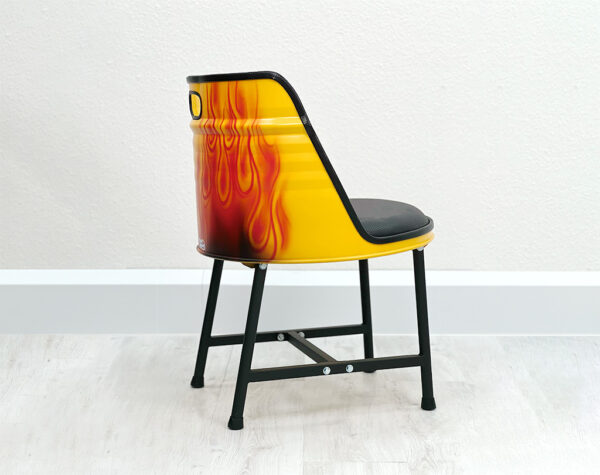 Seitliche Ansicht von Oelfass Esszimmer Stuhl in Gelb mit aufgespraytem Flammendesign auf der Rueckseite, mit schwarzem Sitzpolster und schwarzen Metallbeinen auf weißem Holzboden vor weißem Hintergrund