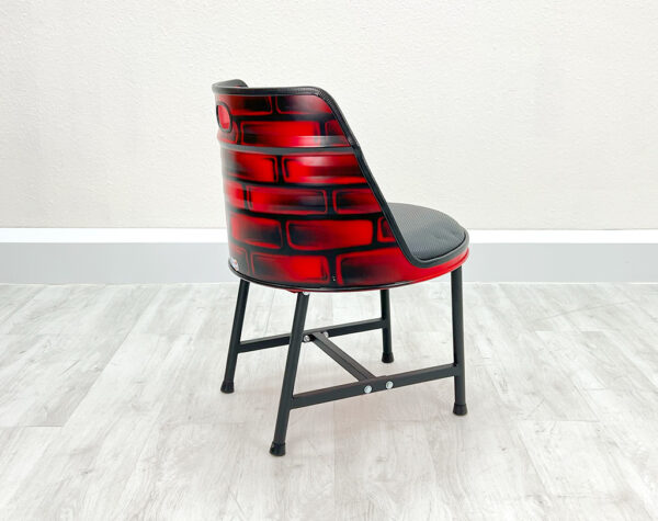 Seitliche Ansicht von Oelfass Esszimmer Stuhl in Rot mit gesprayten Backsteinen auf der Rückseite, mit schwarzem Sitzpolster und schwarzen Metallbeinen auf weißem Holzboden vor weißem Hintergrund