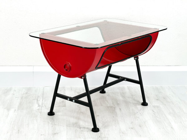 Roter Upcycling Oelfass Tisch mit Stauraum im Inneren und Glastischplatte auf schwarzen Metallfueßen vor weißer Wand auf weißem Holzboden