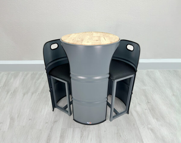 Grauer Stehtisch aus Oelfass mit 2 grauen Fass Barhockern, beides mit dunklem Kantenschutz, mit heller Holztischplatte auf weißem Holzboden vor weißer Wand